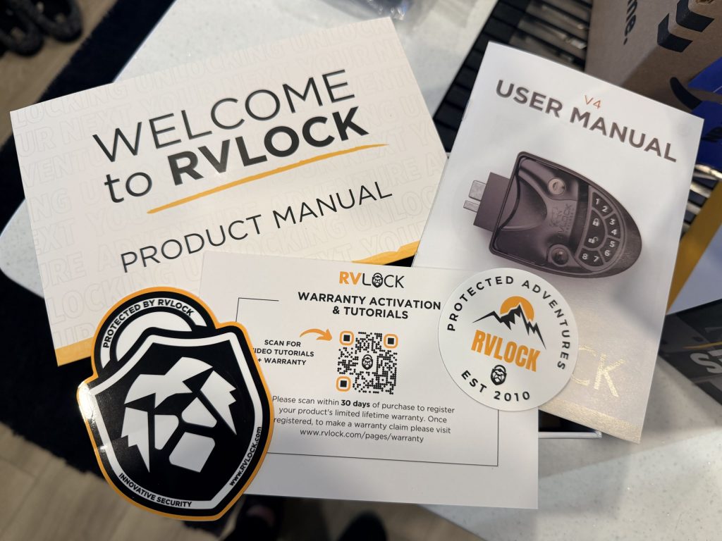 RVLock product materials spread across a countertop including a welcome booklet, V4 user manual, warranty registration card with QR code, and two branded stickers. The printed text includes "WELCOME to RVLOCK PRODUCT MANUAL," "USER MANUAL V4," and "WARRANTY ACTIVATION & TUTORIALS."