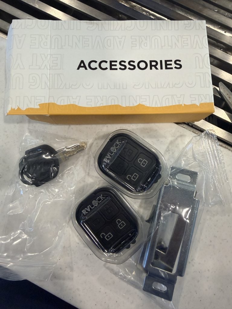 RVLock accessory kit spread out on a countertop with two black key fobs, a backup key, mounting bracket, and a white box labeled "ACCESSORIES." The two remote fobs have lock and unlock buttons and are still sealed in clear plastic packaging.