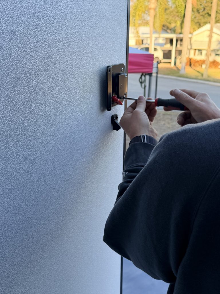 Person removing the original RV door lock with a screwdriver from the inside of a white camper door before installing a new keypad lock. The open doorway looks out onto a campground with palm trees and neighboring RVs in the background.