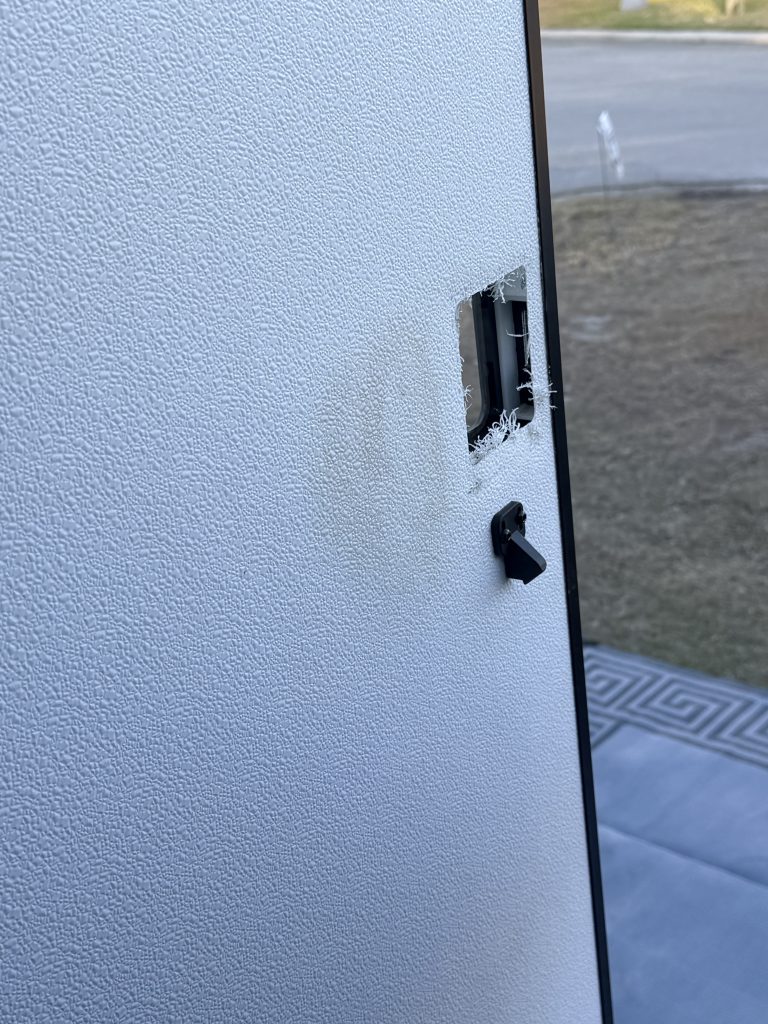 White textured RV door with the original lock removed, leaving a rectangular opening and frayed insulation visible around the cutout. A small black latch remains below the empty opening, with the campsite visible through the open doorway.
