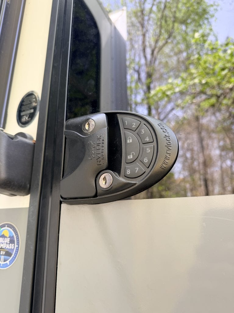 Close up of a black RVLock V4.0 keypad lock mounted on the door of a beige RV. The lock features numbered buttons one through eight and lock and unlock icons, with blurred trees and sunlight reflected in the RV window behind it.