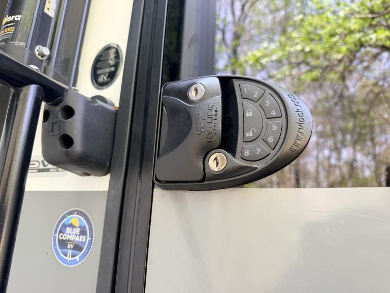 Close up of a black RVLock V4.0 keypad lock mounted on the door of a beige RV. The lock features numbered buttons one through eight and lock and unlock icons, with blurred trees and sunlight reflected in the RV window behind it.