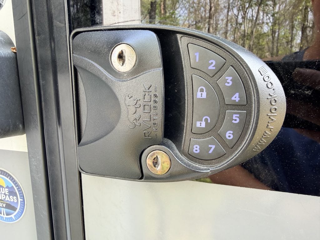 Close up of the RVLock V4.0 keypad installed on an RV door with the number buttons softly glowing blue to show the backlit keypad for nighttime use. The illuminated numbers one through eight and lock and unlock icons are clearly visible against the black lock housing.
