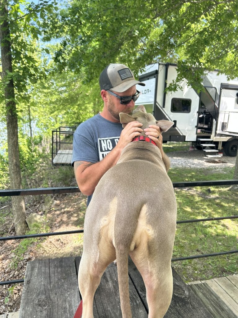 Man wearing sunglasses pets a tan dog standing on a picnic table beside a fifth wheel RV at a shaded campground. The relaxed outdoor setting shows everyday life RVing with dogs while camping under large green trees.