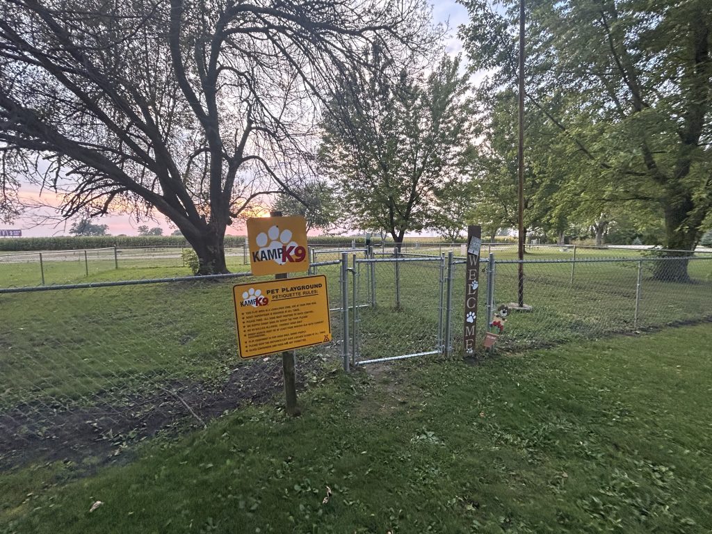 Fenced dog park at an RV campground with signs reading “Kamp K9” and “Pet Playground” near the entrance gate. Trees and open grassy space surround the area, highlighting a pet friendly stop for travelers RVing with dogs.
