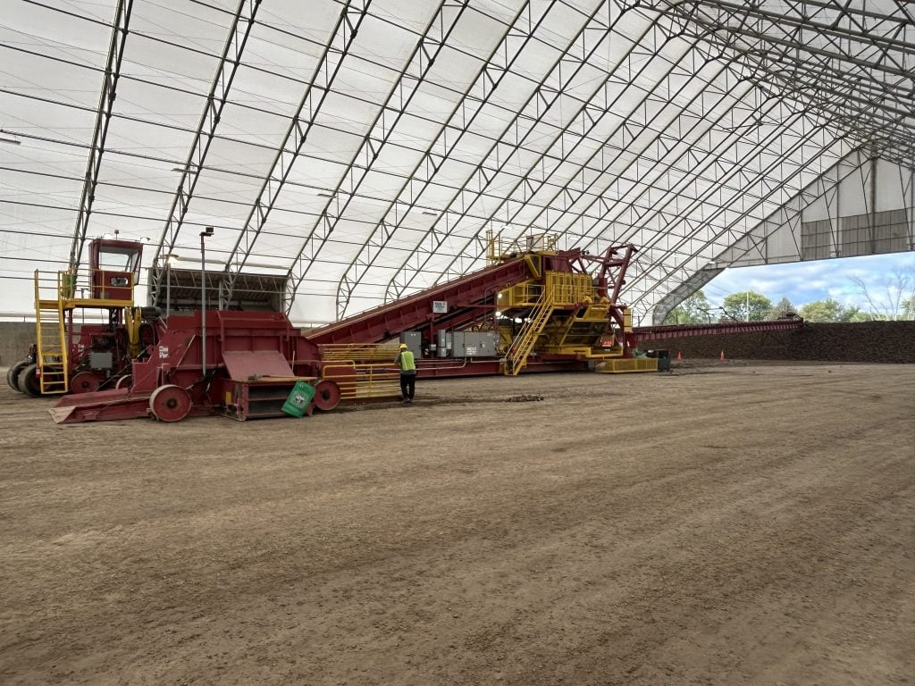 Large red sugar beet processing and conveyor equipment inside a covered agricultural storage facility during the sugar beet harvest. A worker wearing a high visibility vest stands beside the machinery at a seasonal worksite for RVers.