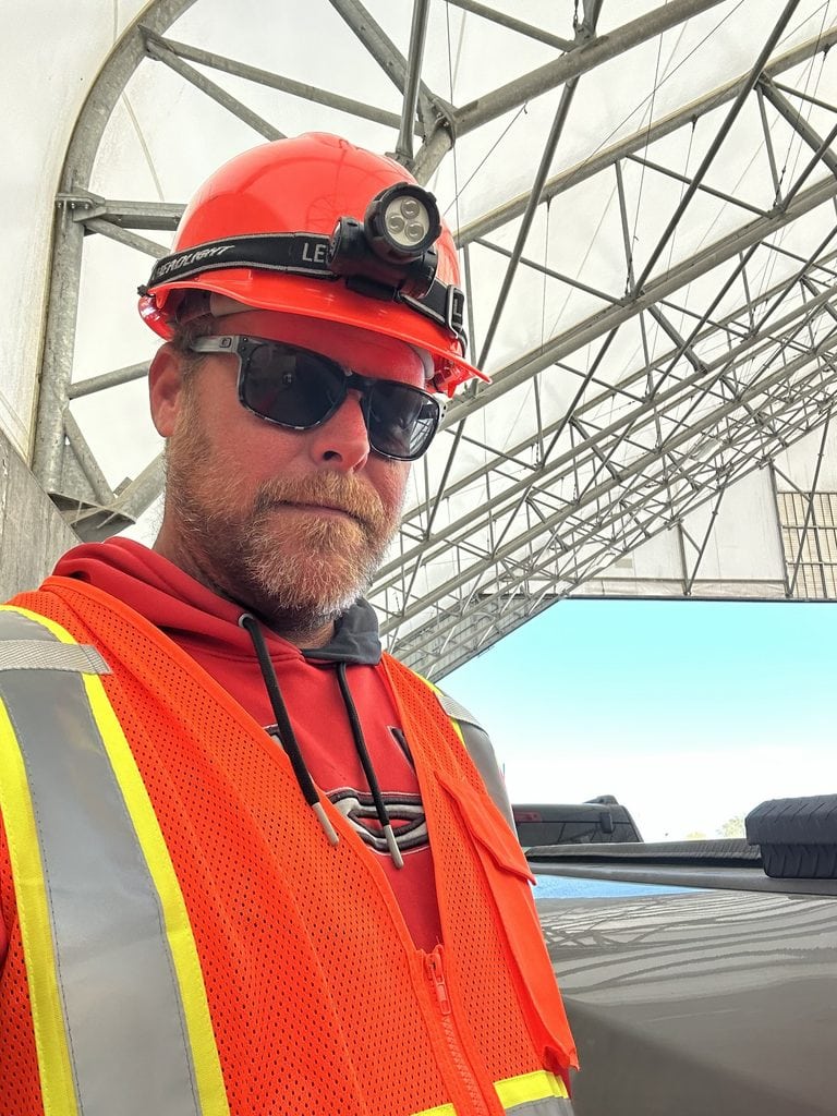 A seasonal sugar beet harvest worker takes a selfie wearing an orange hard hat with a headlamp. He also wears dark safety glasses and a bright orange reflective safety vest while standing beneath the metal framework of a covered agricultural processing facility.