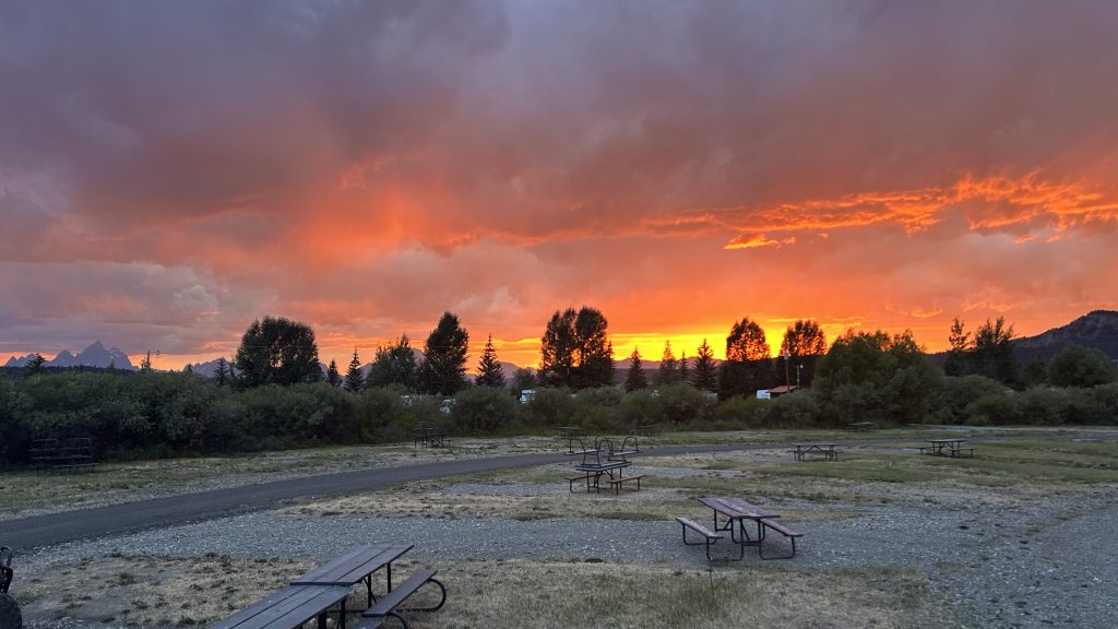A vibrant orange and pink sunset lights up the sky over a quiet workamping site with picnic tables scattered across grassy and gravel campsites. Trees and distant mountain peaks frame the campground beneath dramatic clouds glowing red and purple.
